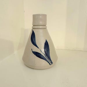 May include: A beige ceramic bottle with a cylindrical neck and a conical body. The bottle is decorated with a blue leaf design. The bottle is sitting on a white surface.