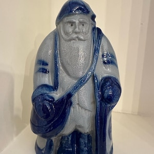 Puede incluir: Figura de cerámica de Papá Noel en tonos azules y grises. La figura lleva un gorro azul, lleva un bolso azul y se apoya en un bastón azul. El Papá Noel tiene una larga barba y bigote blancos. La figura está de pie.