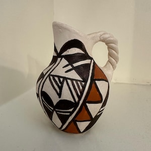 May include: A small, hand-painted ceramic jug with a twisted handle. The jug features a cream-coloured base with black, white, and orange geometric patterns. The spout is cream-coloured, and the handle is a twisted rope design.
