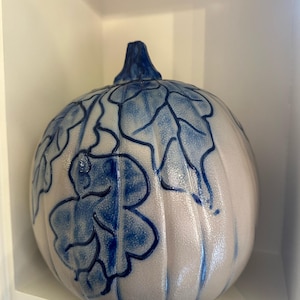 Puede incluir: Una calabaza de cerámica con base beige claro y diseño de hojas azules. La calabaza tiene una superficie texturizada y un tallo azul. El diseño está pintado a mano, agregando un toque único a este artículo de decoración de temporada.