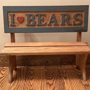 May include: Wooden bench with a backboard that reads "I ❤️ BEARS" in gray letters and a red heart. The bench is made of light brown wood and has a rustic appearance. The bench is suitable for indoor or outdoor use.