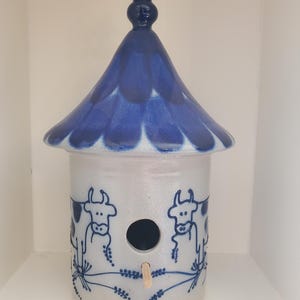 May include: A ceramic birdhouse with a blue roof and finial. The cylindrical body is light grey with blue cow illustrations and floral accents. A round opening is centered below the roof, with a small wooden perch.