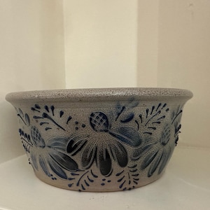 Puede incluir: Un cuenco redondo de gres, con base gris y diseños florales azules. El cuenco tiene un borde ancho y está decorado con flores y hojas estilizadas. Probablemente se utiliza para servir o exhibir.