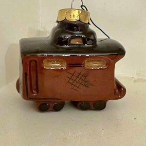May include: A brown ceramic train caboose ornament with a dark brown roof, gold-painted window frames, and a gold metal cap at the top. The side of the caboose features a small, dark, cross-hatched design. A thin green wire loop is attached to the top.