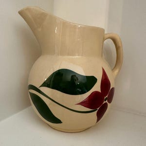 May include: Cream-colored ceramic pitcher with a rounded body and a curved spout. The pitcher is decorated with a large red flower and green leaves. It has a handle on the side.