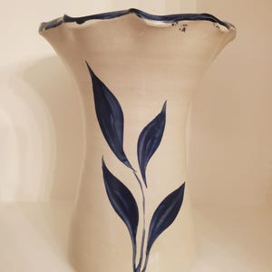 May include: A cream-colored ceramic vase with a flared, ruffled rim and a dark blue trim. The vase features a hand-painted design of dark blue leaves. The vase has a wide opening and a rounded body, suitable for displaying flowers.