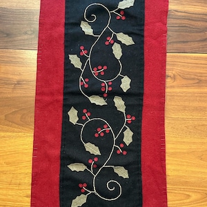 May include: A rectangular, festive table runner with a red border and a black center. The center features an embroidered design of holly leaves and red berries. The runner is made of felt and is suitable for holiday decor.