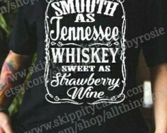 Può includere: T-shirt nero con un design grafico bianco. Il design recita "Smooth as Tennessee Whiskey, Sweet as Strawberry Wine" in stile vintage. La maglietta è a girocollo.