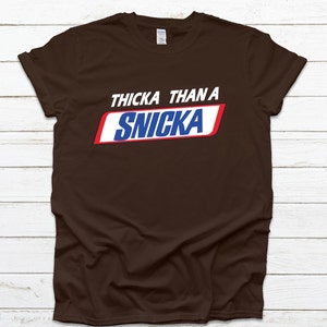 May include: A brown t-shirt featuring the text "THICKA THAN A SNICKA" in white, red, and blue, styled after the Snickers logo. The garment is presented against a white wooden backdrop.