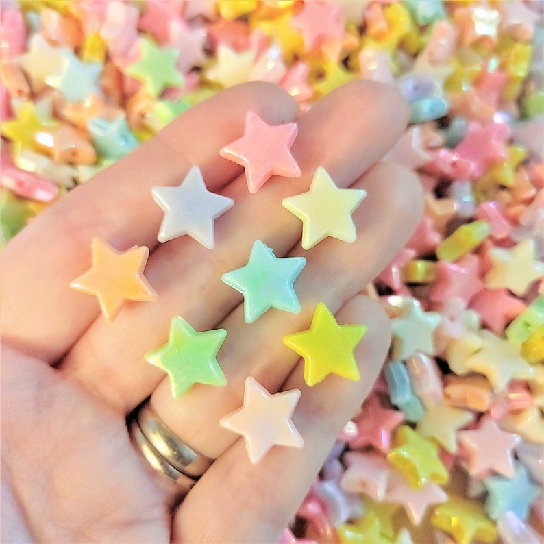 K&ouml;nnte beinhalten: Eine Hand h&auml;lt eine Vielzahl von pastellfarbenen sternf&ouml;rmigen Perlen. Die Perlen sind aus Kunststoff und haben eine gl&auml;nzende Oberfl&auml;che. Die Perlen sind in einem zuf&auml;lligen Muster angeordnet.