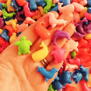May include: A collection of colorful plastic sea animal beads, including whales, dolphins, turtles, and fish. The beads are arranged in a hand, showcasing their vibrant colors and playful designs.