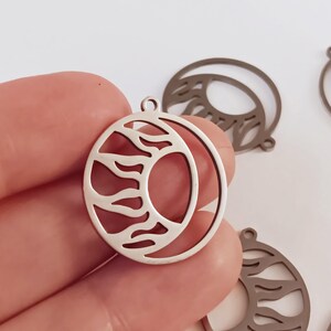 May include: Silver metal charm with a sun and moon design. The sun is depicted as a stylized flame and the moon is a crescent shape.