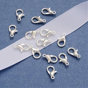 May include: Silver-toned lobster clasps, a type of jewelry finding, are scattered on a white surface. These clasps are used for attaching jewelry chains or cords to other jewelry pieces.
