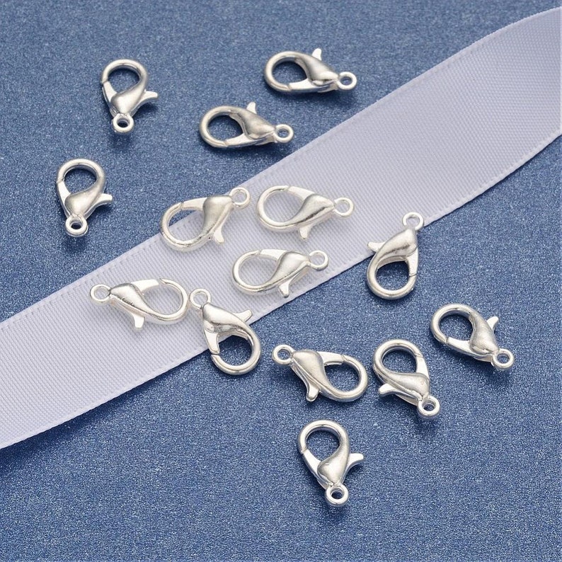 May include: Silver-toned lobster clasps, a type of jewelry finding, are scattered on a white surface. These clasps are used for attaching jewelry chains or cords to other jewelry pieces.