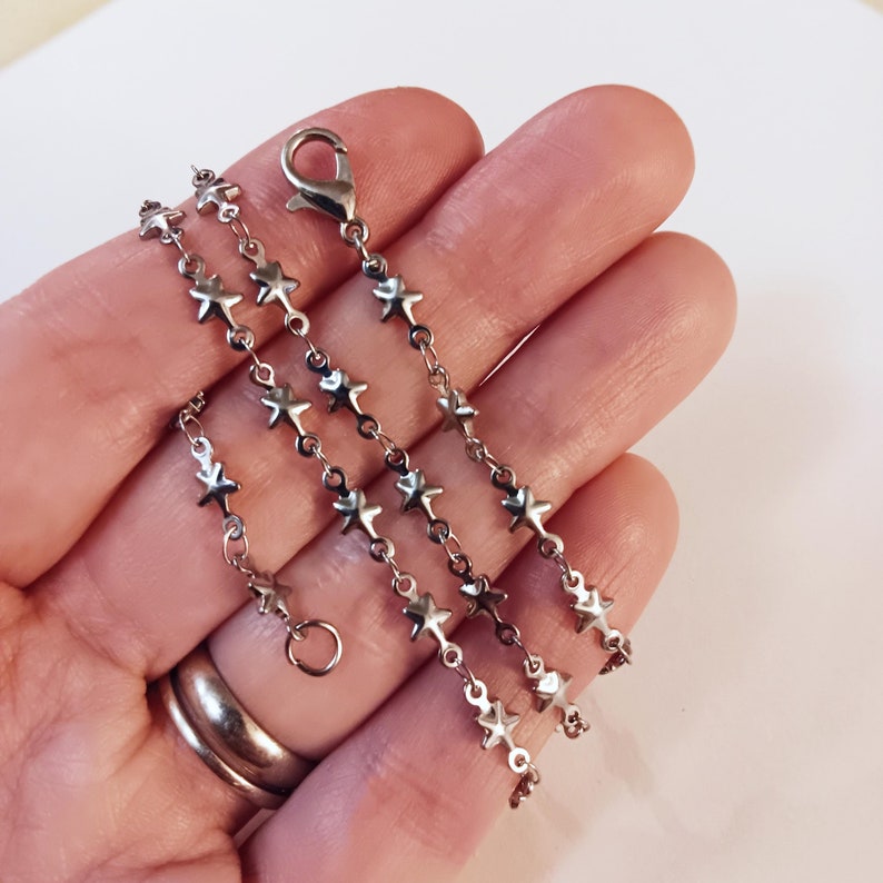 May include: A silver chain necklace with star-shaped links. The chain is layered on a hand, showing the length and detail of the necklace.