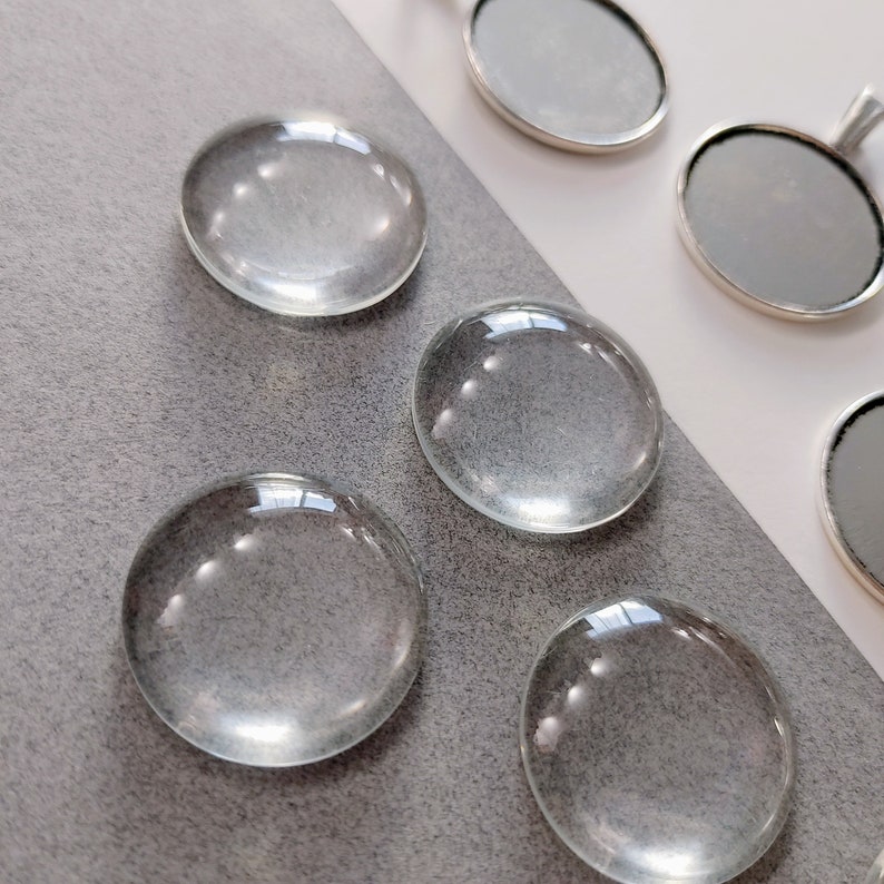 May include: Five clear glass cabochons, round and smooth, with a slight dome shape. The cabochons are arranged in a row on a grey surface.