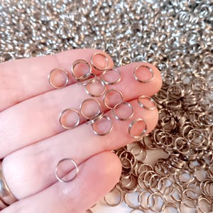 May include: Close-up of small, silver-colored jump rings scattered on a surface and held in a hand. The rings are circular and appear to be made of metal. The background is filled with more jump rings, suggesting a craft or jewelry-making theme.