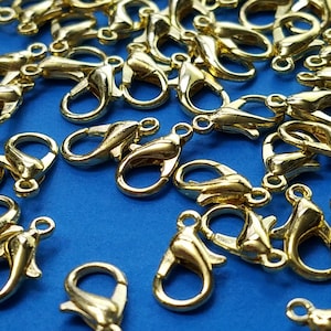 May include: A pile of gold-colored metal lobster claw clasps on a blue background. These clasps are used for jewelry making and crafting.