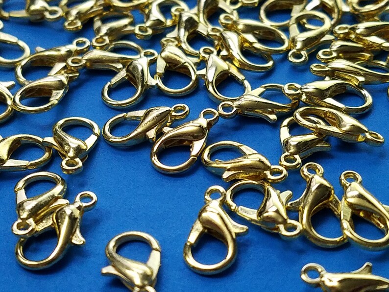 May include: A pile of gold-colored metal lobster claw clasps on a blue background. These clasps are used for jewelry making and crafting.