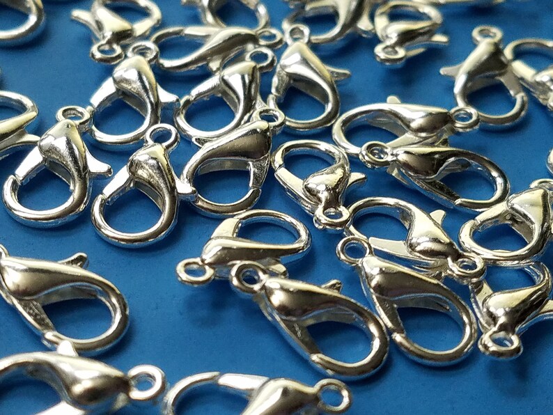 May include: A close-up of a group of silver-toned lobster claw clasps. The clasps are scattered on a blue surface.