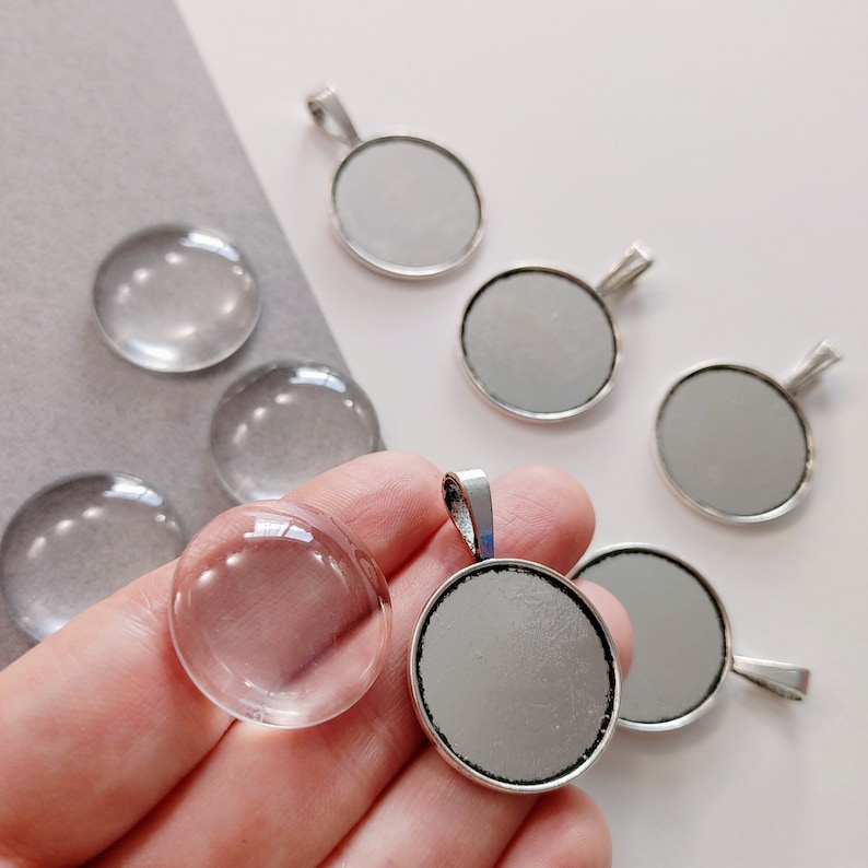 May include: A set of eight round glass and metal pendant trays. The trays are silver with a smooth, reflective surface. The glass is clear and has a slightly domed shape. The trays are ideal for creating custom jewellery or keychains.