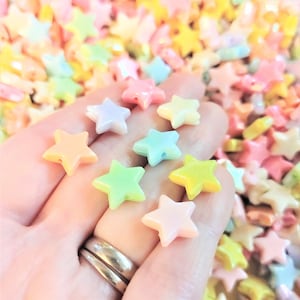 K&ouml;nnte beinhalten: Nahaufnahme einer Hand, die eine Vielzahl von pastellfarbenen sternf&ouml;rmigen Perlen h&auml;lt. Die Perlen sind aus Kunststoff und haben eine gl&auml;nzende Oberfl&auml;che.