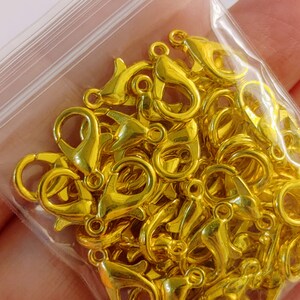 May include: A close-up of a bag of gold-colored metal lobster clasps. The clasps are small and have a shiny finish.