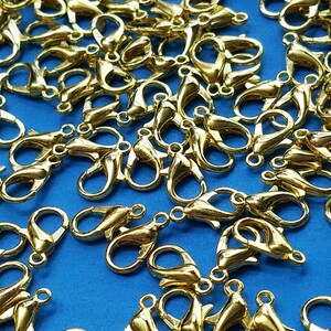 May include: A large group of gold-colored metal lobster claw clasps, scattered on a blue surface. These clasps are commonly used for jewelry making and crafting.