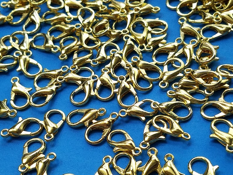 May include: A large group of gold-colored metal lobster claw clasps, scattered on a blue surface. These clasps are commonly used for jewelry making and crafting.