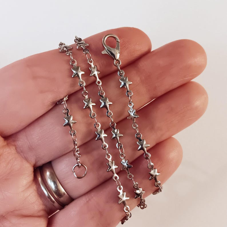 May include: A silver chain necklace with star-shaped links. The chain is shown on a hand, with the clasp visible.