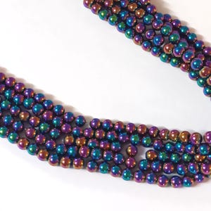 Op de afbeelding: Een streng iriserende hematiet kralen. De kralen zijn rond en hebben een metallic afwerking met tinten van paars, blauw, groen en goud.