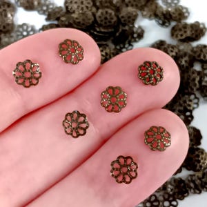 May include: Close-up of several small, bronze-coloured filigree flower-shaped beads. The beads have an openwork design with a red centre. Some beads are in focus, while others are blurred in the background. These beads are ideal for jewellery making and crafting.