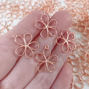 May include: Close-up of four rose gold flower charms with a loop at the top for jewelry making. The charms are in focus, with other charms slightly blurred in the background. The charms are delicate and lightweight.