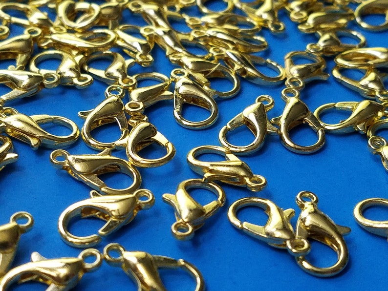 May include: A close-up of a group of gold-toned metal lobster claw clasps. The clasps are scattered on a blue surface.