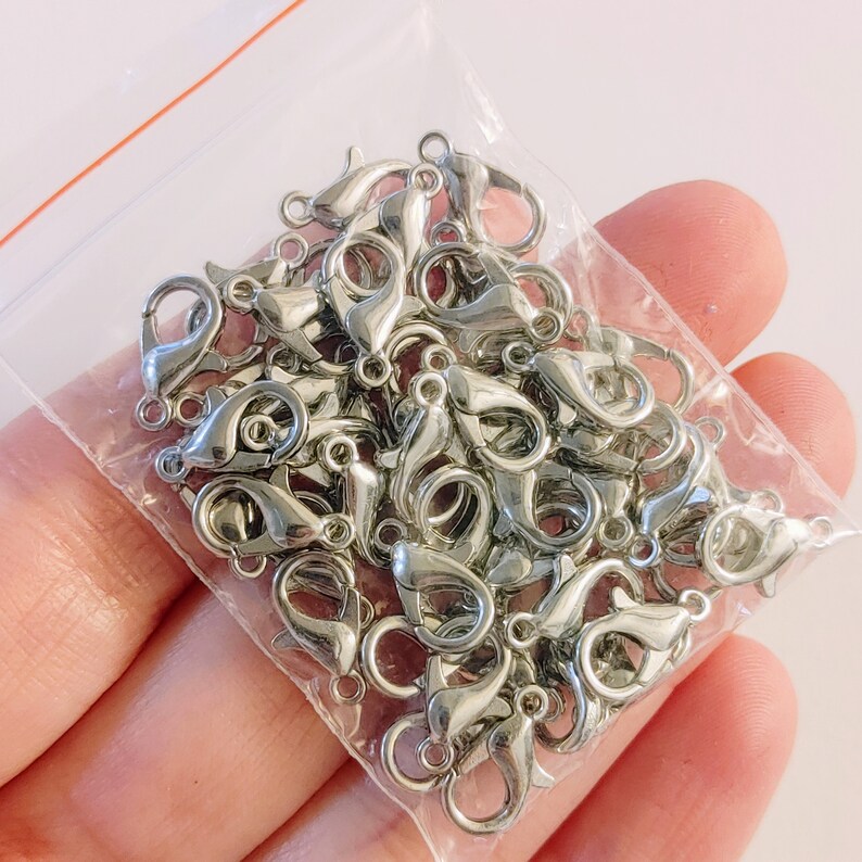 May include: A close-up of a bag of silver lobster claw clasps. The clasps are arranged in a pile and are ready to be used for jewelry making.