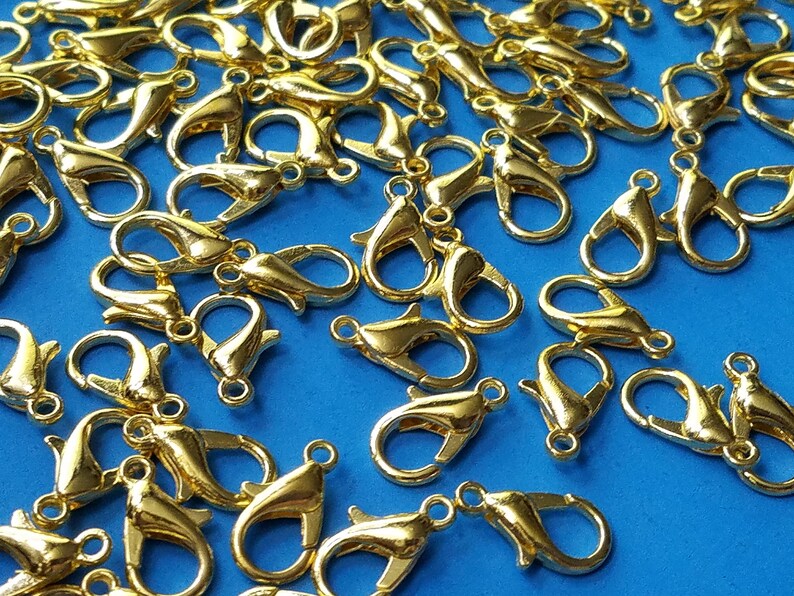 May include: A large group of gold-colored metal lobster claw clasps. The clasps are scattered on a blue surface.