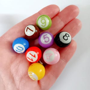 May include: A collection of miniature billiard balls in various colors, including blue, red, and yellow, each with a different number. The balls are held in a hand, showcasing their small size and vibrant appearance. The numbers are in white.