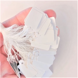 May include: A bunch of white blank gift tags with string attached. The tags are square shaped and have a hole punched in the top corner.