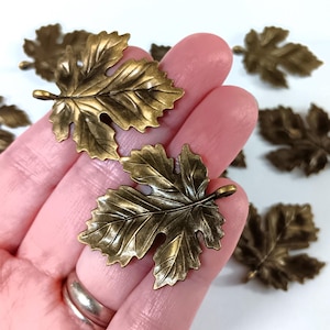 May include: Close-up of several bronze-colored leaf charms. The charms are detailed with veining and have a small loop at the stem. The leaves are scattered on a white surface, with some held in a hand.