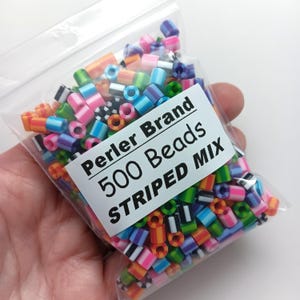 Puede incluir: Una bolsa de pl&aacute;stico transparente llena de cuentas Perler a rayas de colores. La bolsa tiene una etiqueta blanca que dice "Perler Brand 500 Beads STRIPED MIX". Las cuentas son una mezcla de rayas rosas, azules, naranjas, verdes y blancas y negras.