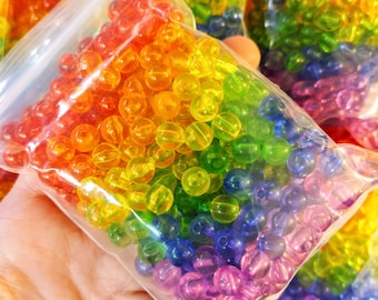 May include: A clear plastic bag filled with rainbow coloured beads. The beads are round and translucent, and they are arranged in a rainbow pattern. The bag is open at the top, and the beads are spilling out.