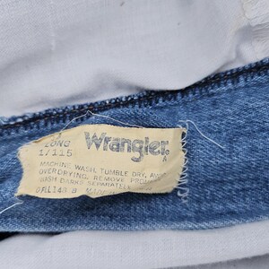 May include: Close-up of a blue denim Wrangler jeans label with white stitching. The label reads "Long 1/115" and includes washing instructions.