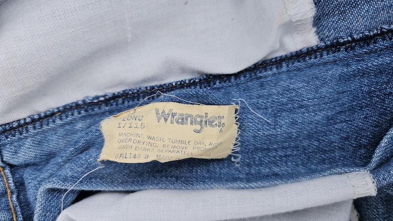May include: Close-up of a blue denim Wrangler jeans label with white stitching. The label reads "Long 1/115" and includes washing instructions.