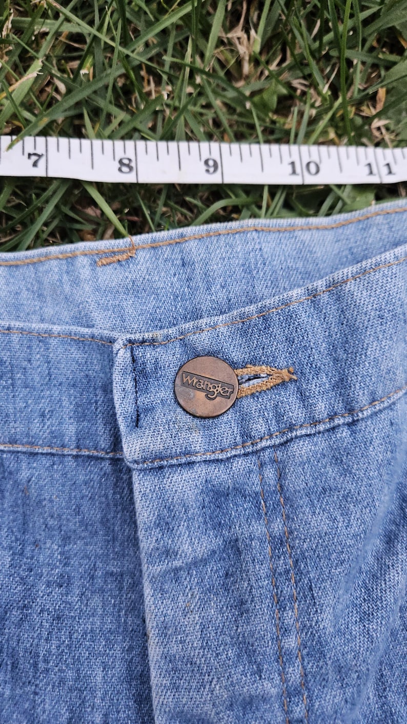 May include: Close-up of a blue denim jean button with a brown metal Wrangler button.