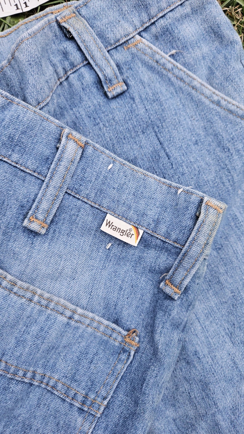 May include: A pair of light blue denim jeans with a brown leather Wrangler label. The jeans have a classic five-pocket style and are folded over.