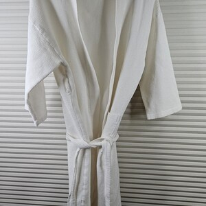 May include: A white waffle-weave robe with a matching belt, hanging on a wooden hanger. The robe has a shawl collar and long sleeves. The background is a wall with horizontal gray and white stripes.