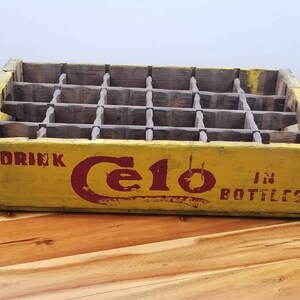 May include: A vintage wooden crate for holding bottles of Cel-O drink. The crate is yellow with red lettering that says "Drink Cel-O in Bottles".