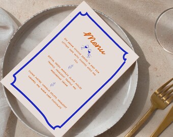 May include: A menu card with a blue border and the word "Menu" in orange script. The menu lists dishes like sea bream ceviche, truffle pasta, and choux pastry. The card is on a white plate with gold cutlery.