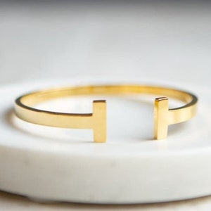 Op de afbeelding: Een goudkleurige armband met een T-vormig ontwerp, rustend op een witte, cirkelvormige schaal. De armband heeft een minimalistische stijl en een gepolijste afwerking. De achtergrond is zachtgrijs, wat een contrast creëert met het sieraad.