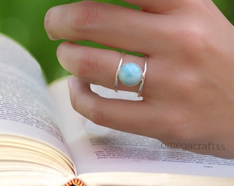 May include: A silver ring with a light blue gemstone set in a double band design. The ring is worn on a hand holding a book.